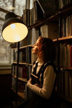 a woman sitting in front of a book shelf next to a lamp