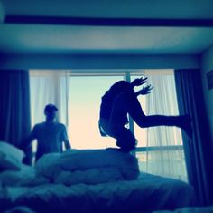 a person jumping on top of a bed in front of a window and another person looking out the window