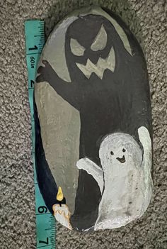 a painted rock with a ghost and cat on it next to a measuring tape in the shape of a jack - o - lantern