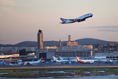 an airplane is flying over the airport with other planes on the tarmac and buildings in the background