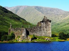 an old castle sitting on the shore of a lake in front of mountains and hills