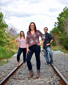 three people standing on train tracks in front of trees and bushes, posing for the camera