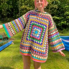 a woman with pink hair wearing a colorful crocheted sweater and shorts in the grass