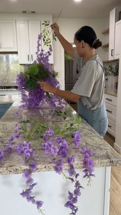 a woman arranging purple flowers on top of a kitchen counter