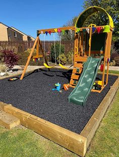 a wooden play set with a green slide and black gravel in the ground next to it