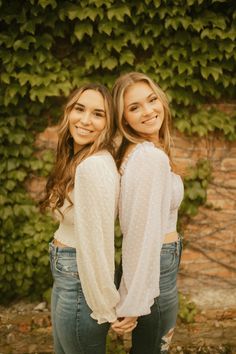 two young women standing next to each other in front of a green plant covered wall