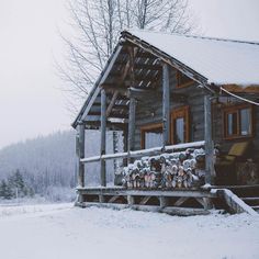 a log cabin in the snow with logs stacked on it's porch and windows