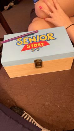 a woman is sitting on the floor with her hands in her mouth and holding a box that says senior story