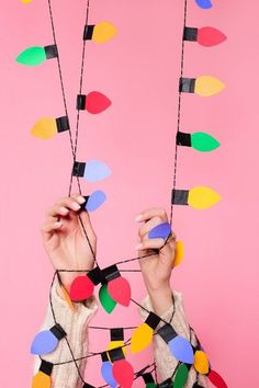 a woman is holding up some string with colored lights attached to her hands on a pink background