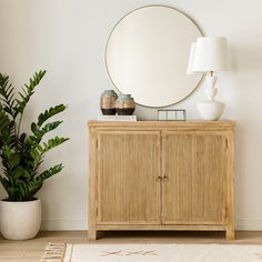 a wooden cabinet sitting next to a plant and a large round mirror on the wall