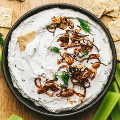 a bowl filled with white dip surrounded by celery and crackers on a wooden table