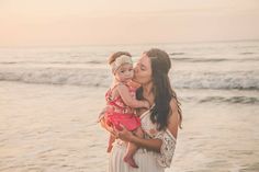 a woman holding a baby in her arms while standing on top of the beach next to the ocean