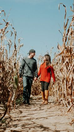 a man and woman walking down a dirt path through a corn field holding hands, with the sky in the background