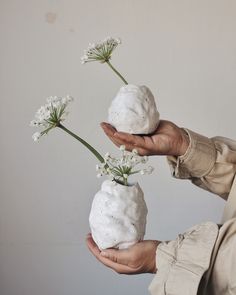 a person holding two white vases with flowers in them on their hands, one is made out of clay