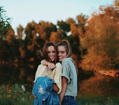 two young women hugging each other in front of trees and grass with fall foliage behind them