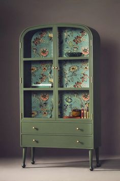 a green cabinet with floral wallpaper on the back and shelves below it, in front of a gray background