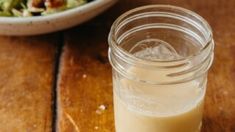 a glass filled with liquid sitting on top of a wooden table next to a bowl of salad