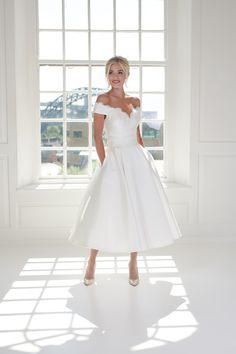 a woman standing in front of a window wearing a white dress with off the shoulder sleeves