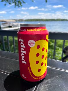 a can of soda sitting on top of a wooden table next to the ocean and trees