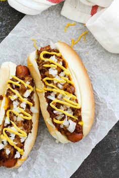 two chili dogs with mustard and onions on them sitting on wax paper next to napkins