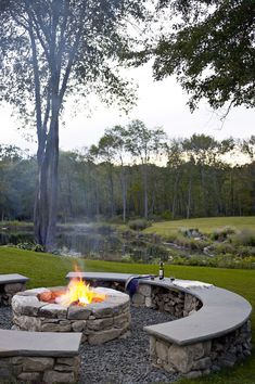 a fire pit in the middle of a grassy area with trees and water behind it