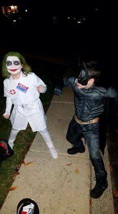 two kids dressed up as batman and robin wayne on the sidewalk at night time with their luggage