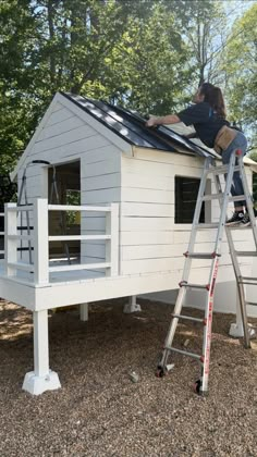 a man standing on top of a ladder next to a small white house in the woods