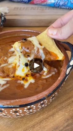 a hand holding a tortilla chip over a bowl of chili cheese and beans