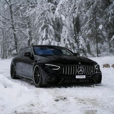 a black car driving down a snow covered road in front of some trees and bushes