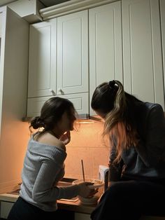 two women sitting at a kitchen counter in front of a stove top oven, one holding a knife and the other writing on a piece of paper