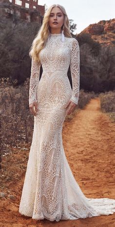 a woman standing on a dirt road wearing a long sleeved wedding dress with an open back