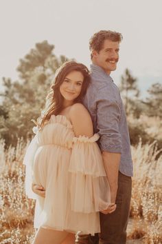 a man and woman standing next to each other in tall grass with trees behind them