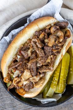 a close up of a hot dog in a pan with pickles and mushrooms on the side