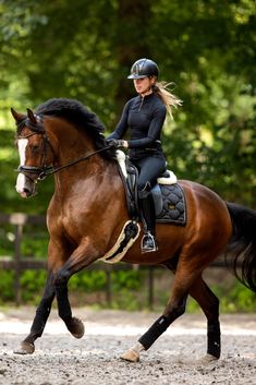 a woman riding on the back of a brown horse through a dirt field with trees in the background