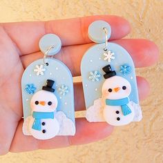 a hand holding two snowman earrings in it's left hand, one is wearing a hat and the other has a blue scarf