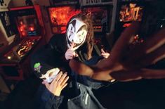 two people with masks on in front of some pinball machines and one person holding up a mask