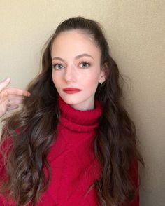 a woman with long dark hair wearing a red turtle neck sweater and holding her hand up in the air