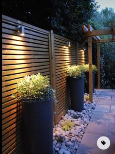 some plants are in large black pots on the side of a wooden fence at night