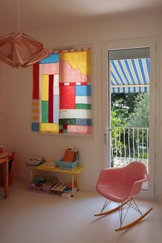 a pink rocking chair in front of a window with colorful quilts hanging on it