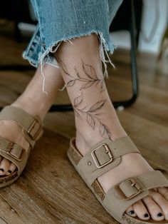 a woman's legs with tattoos on them and her feet in sandals, sitting on a wooden floor