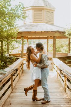 a man and woman standing on a wooden bridge in front of a gazebo with their arms around each other