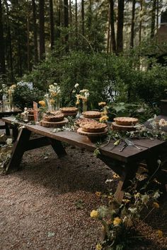 a picnic table with pies and flowers on it in the middle of a forest