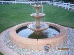 a water fountain in the middle of a brick circle with grass and white fence behind it