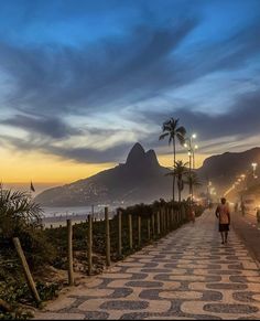 a man walking down a street next to the ocean at dusk with mountains in the background