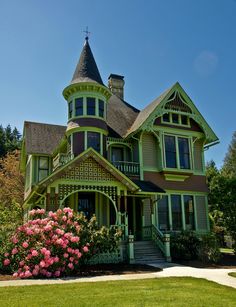 a green victorian style house with pink flowers in the foreground and trees in the background