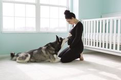 a woman kneeling down next to a dog on the floor in front of a crib