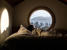 a person laying in bed with their head on the pillow and looking out an oval window