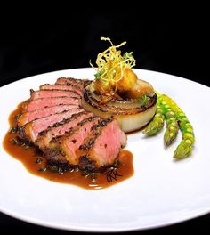 a white plate topped with meat and vegetables on top of a sauce covered table next to asparagus