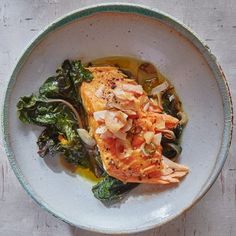 a white bowl filled with fish and greens on top of a wooden table next to utensils