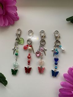 four different colored butterfly charms on a white surface next to pink flowers and a purple flower